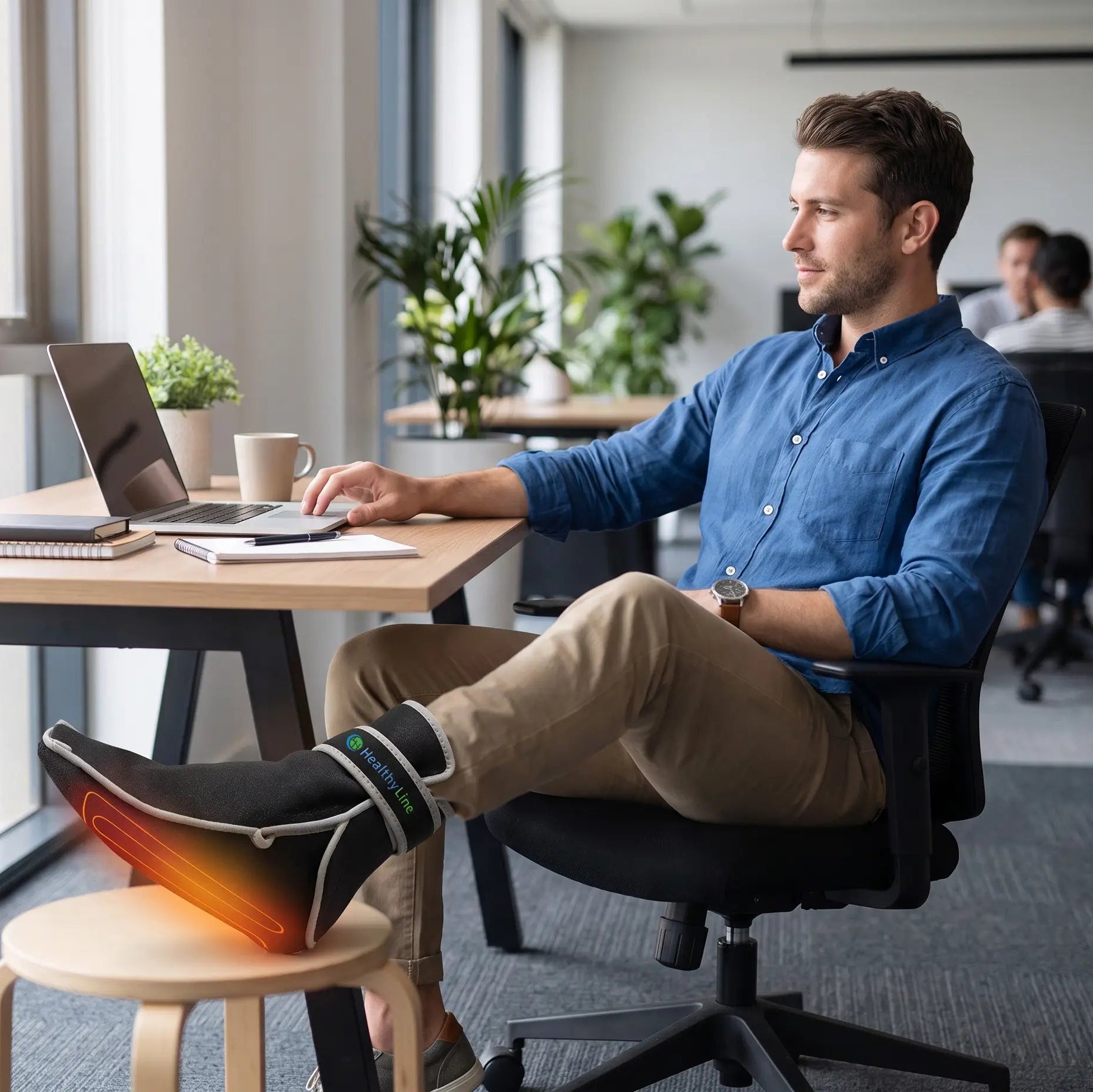 Man working at office desk with foot propped up wearing a HealthyLine heated foot wrap.
