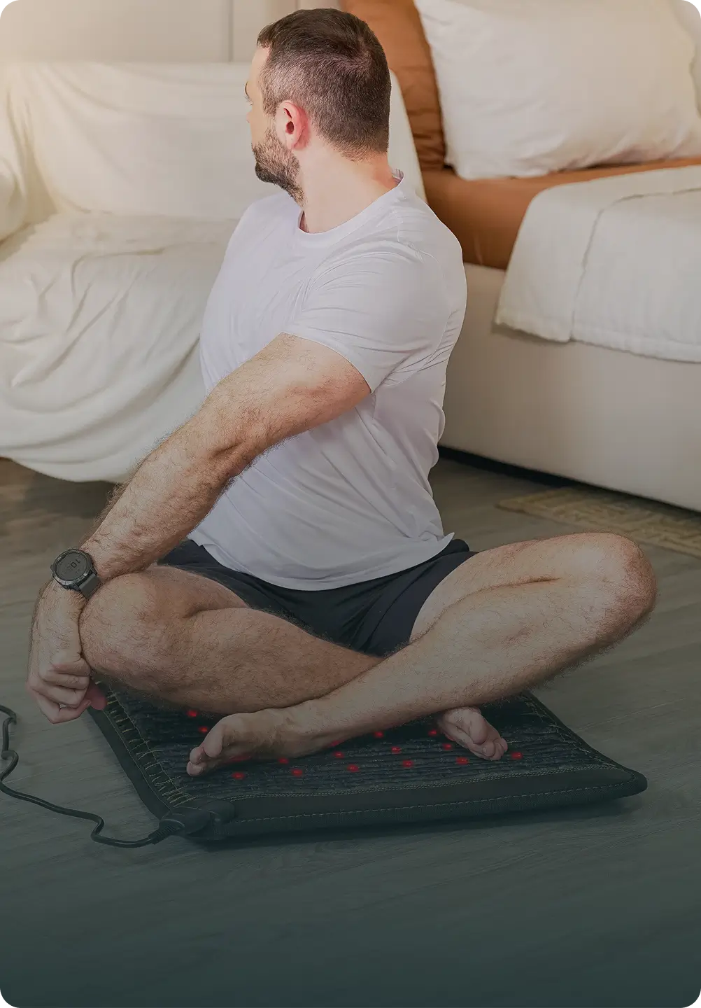 Man seated cross-legged on the HealthyLine Jet Mat 4020 with red infrared LEDs active, next to a bed