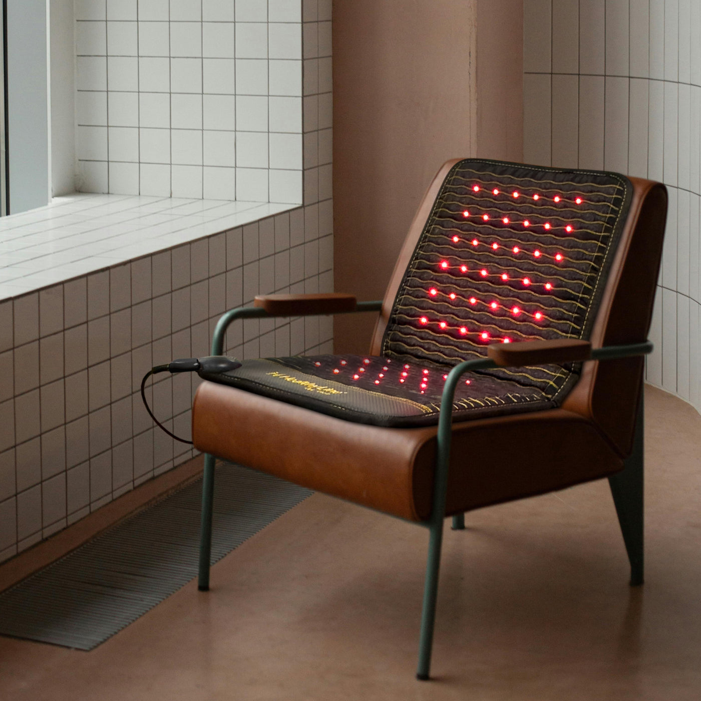 Jet Chair mat placed on a brown leather mid-century style chair with green metal legs in a room with white tile walls, red photon lights glowing on the seat and back