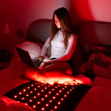 Woman sitting cross-legged on a bed using a laptop, with the Jet 72x24 inch mat glowing red beneath her in a dim room lit by red light therapy