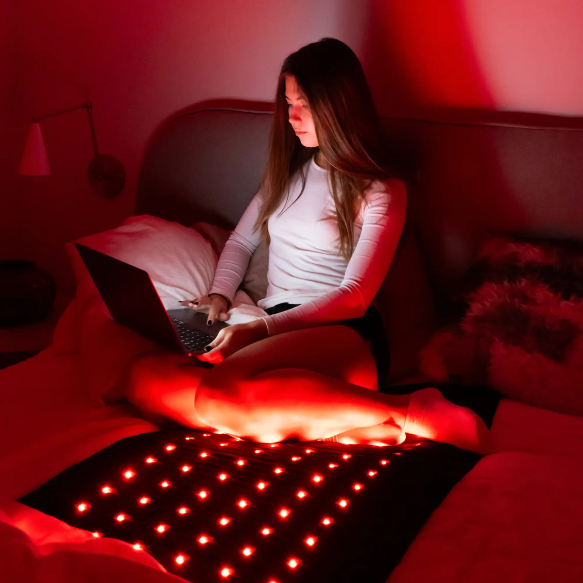 Woman sitting on a bed with a laptop, legs resting on the glowing HealthyLine Jet Mat 7428 in a dimly lit room
