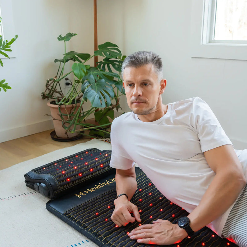 Man seated on the HealthyLine Jet Mat 7224 with the Jet Pillow beside him, both showing active red infrared LEDs