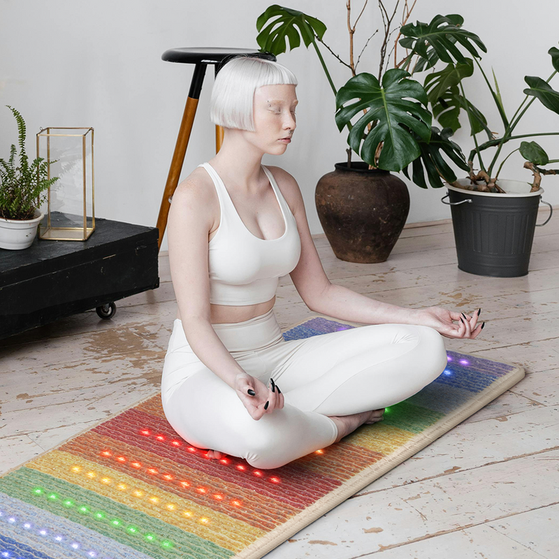 Woman in a white robe meditating on a HealthyLine Rainbow Chakra 74x28 mat with glowing colorful photon lights