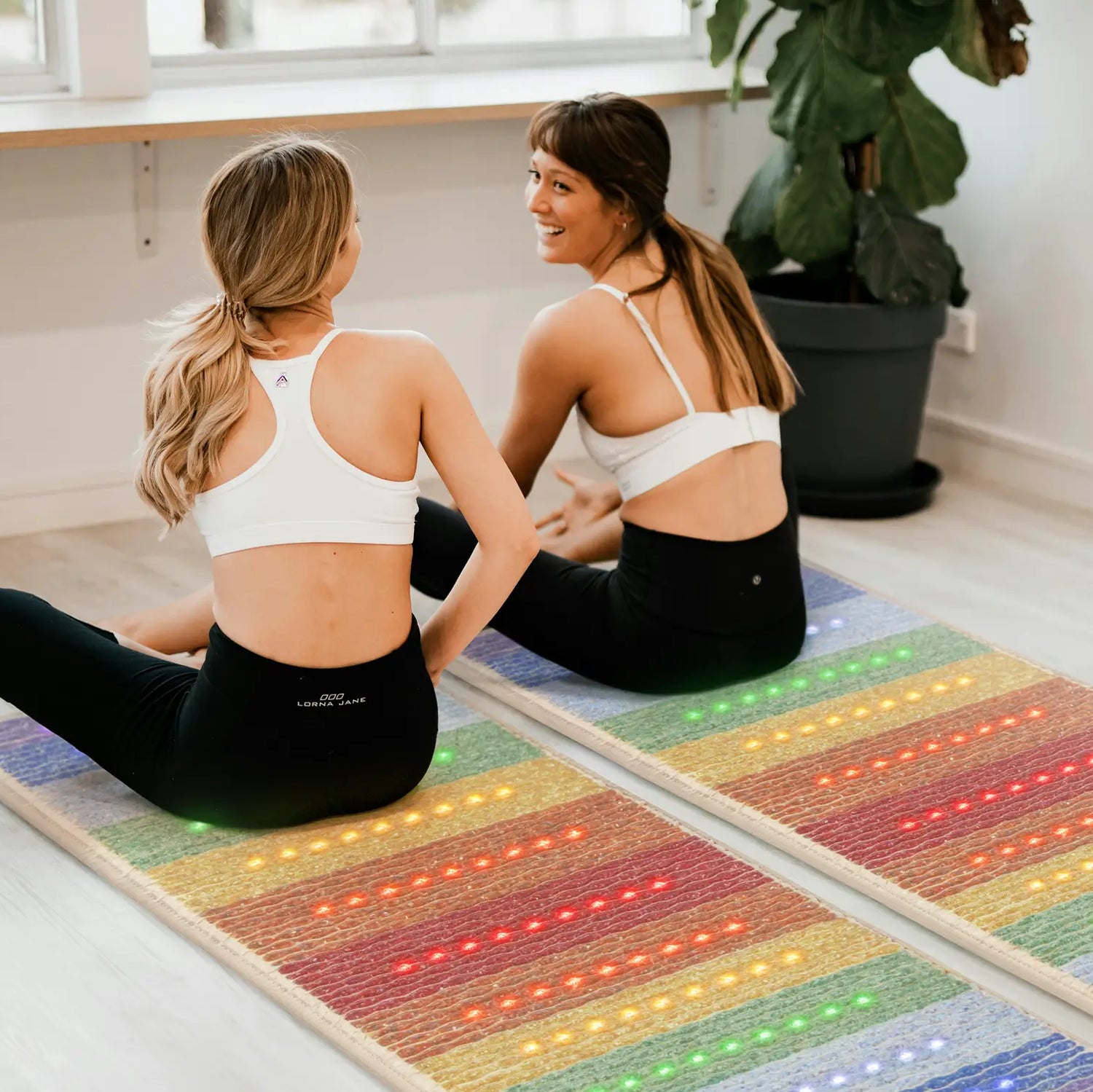Two women sitting on HealthyLine Rainbow Chakra 7428 infrared mats with glowing multicolor photon lights