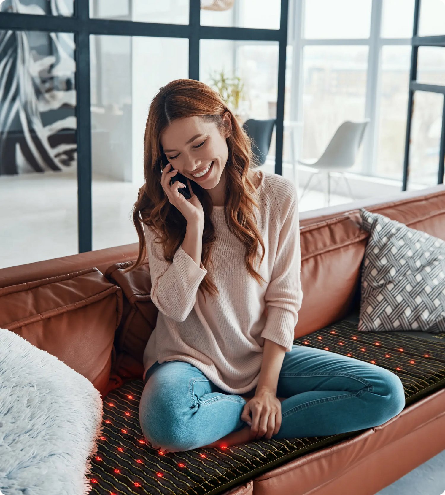 Woman smiling while talking on phone, seated cross-legged on a HealthyLine Jet 74x28 infrared therapy mat