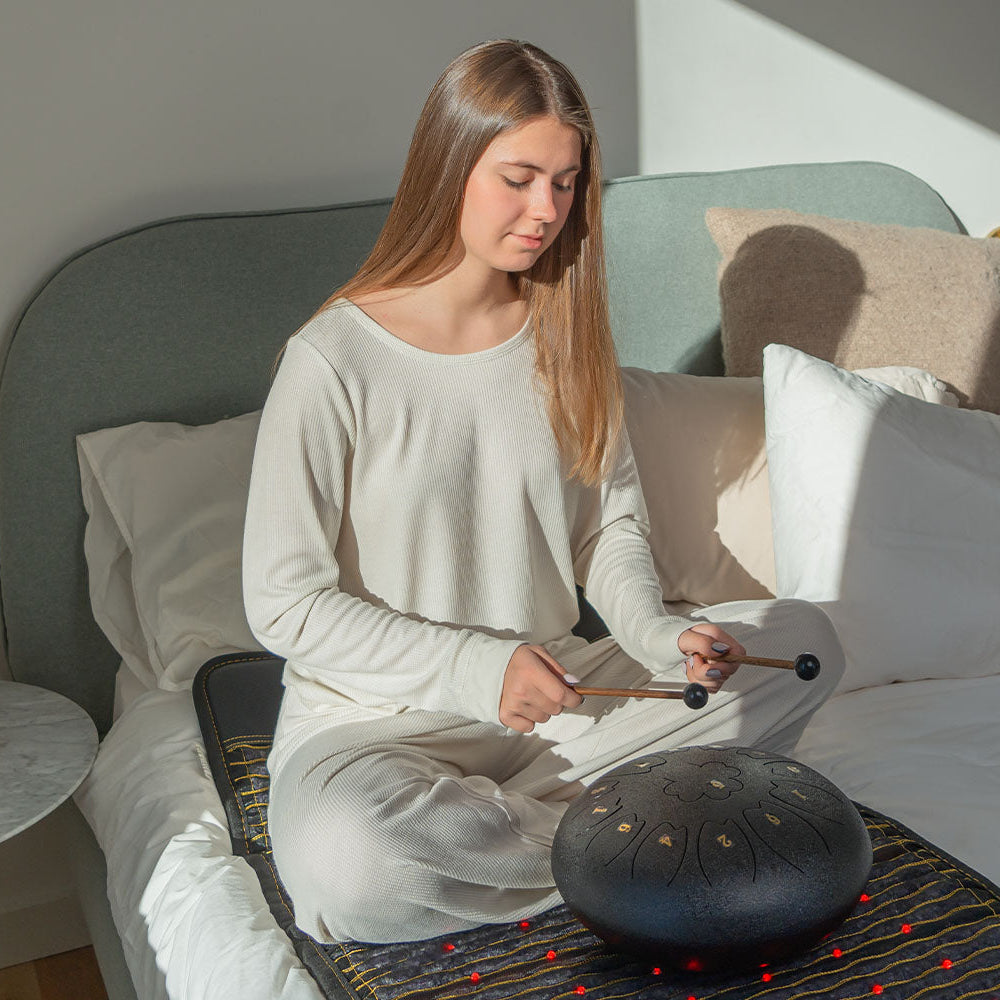 Woman sitting on a HealthyLine Jet 72x24 PEMF mat on a bed, playing a steel tongue drum with mallets