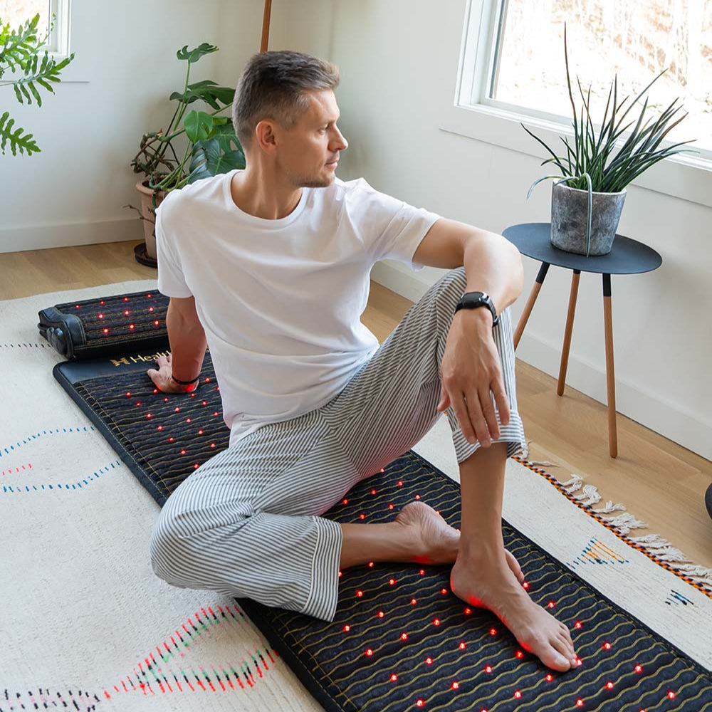 Man sitting cross-legged on a HealthyLine infrared Jet 72x24 mat with red light LEDs illuminated, in a bright living room