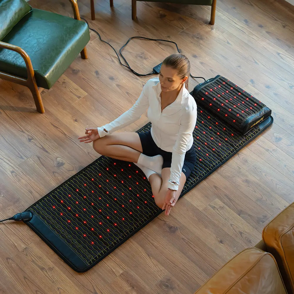Woman sitting cross-legged in meditation pose on the Jet 72x24 mat with red photon lights glowing on a wood floor