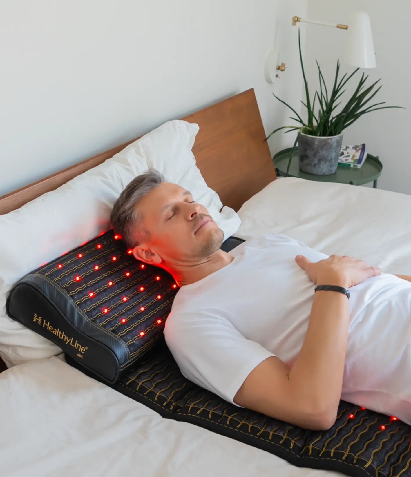 Man sleeping on his back with a HealthyLine Jet mat and pillow supporting his neck, red LEDs glowing.