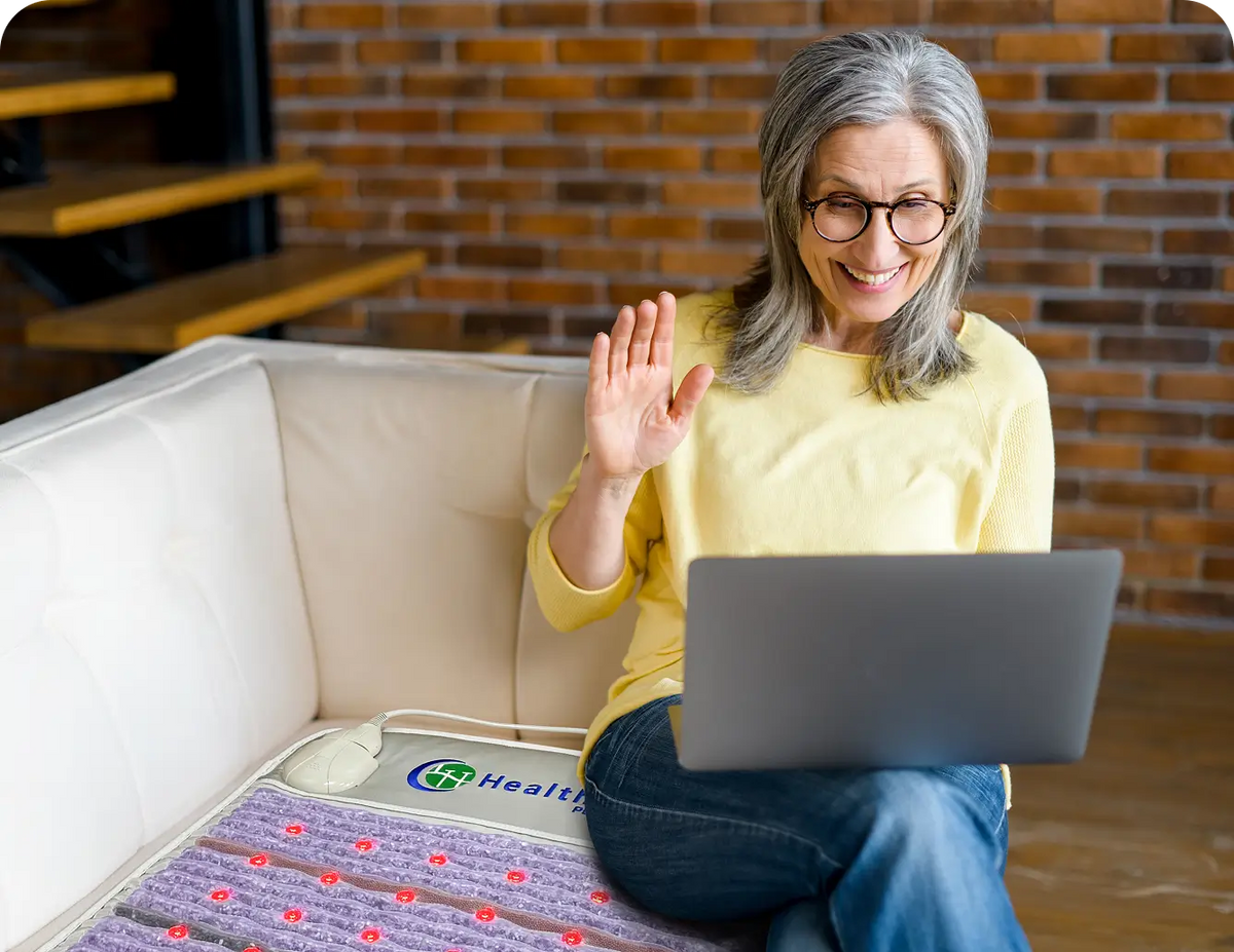 Woman video chatting on a laptop while seated on a sofa next to the HealthyLine Platinum Mat 7224 with red infrared LEDs