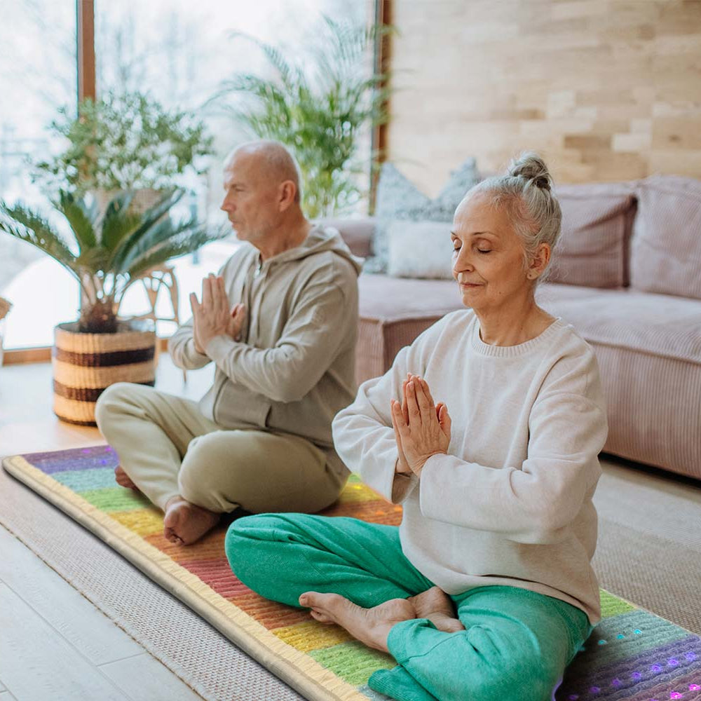Two older adults meditating in prayer pose on a Rainbow Chakra 7428 PEMF mat in a sunlit living room