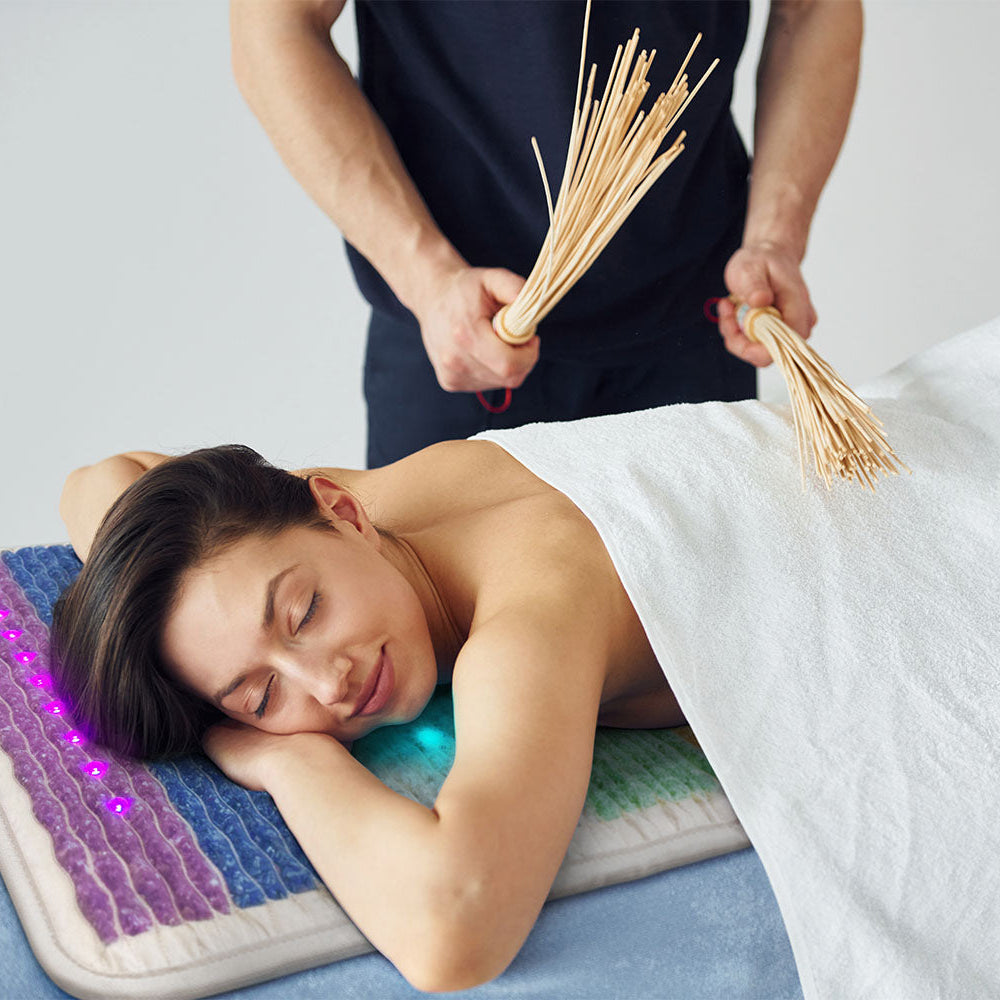 Woman lying face-down on a Rainbow Chakra mat with glowing purple LEDs while a therapist performs a reed massage