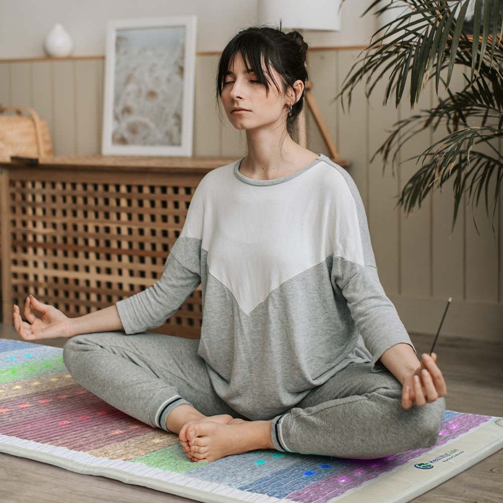 Woman meditating cross-legged with incense on a HealthyLine Rainbow Chakra mat with glowing multicolor photon lights