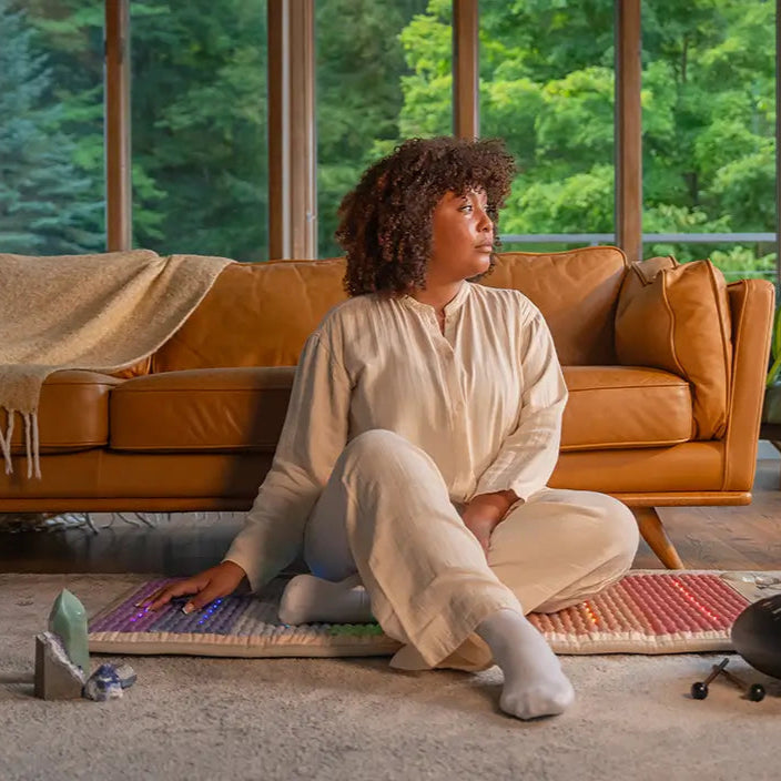 
                  
                    Person sitting cross-legged on Rainbow Chakra 50x24 mat, 7 chakra color bands visible beneath them
                  
                