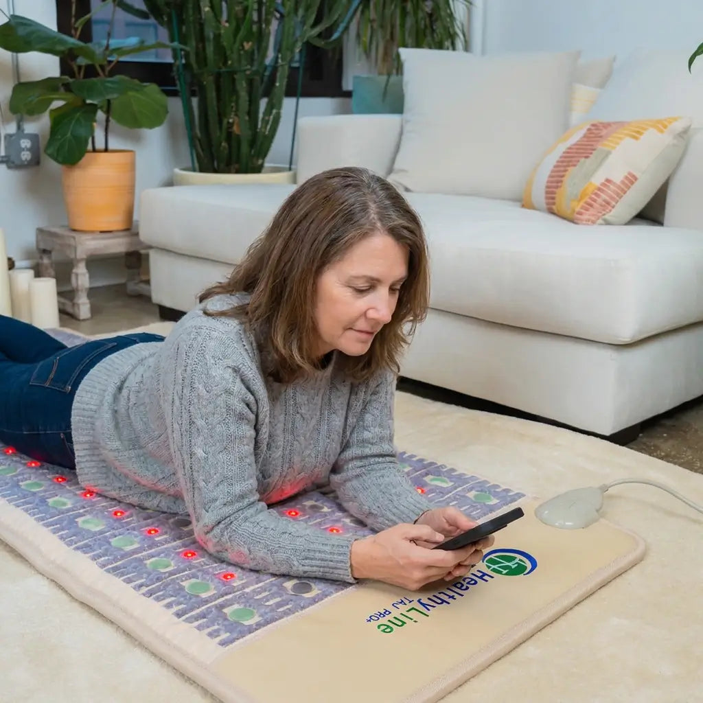 Woman lies on her stomach on a HealthyLine TAJ Mat 7428 with glowing red photon lights, looking at her phone
