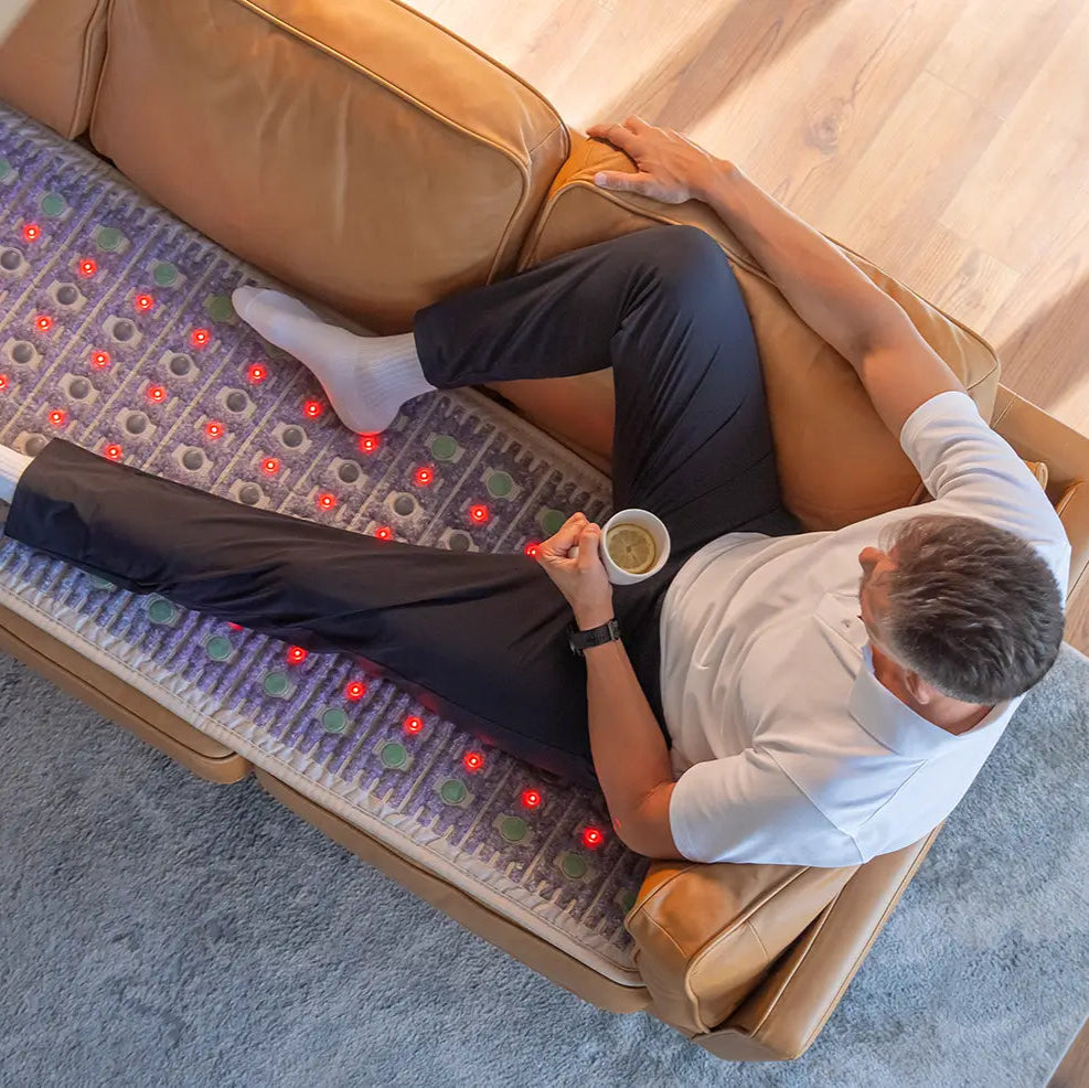 Man lying on a leather sofa holding a coffee cup, resting on the TAJ 72x24 mat with red photon lights glowing beneath him