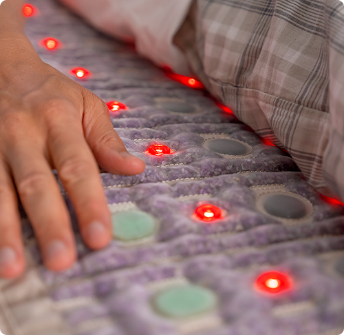 Close-up of a hand on a HealthyLine TAJ mat with glowing red infrared LEDs and embedded amethyst, jade, and tourmaline gemstones