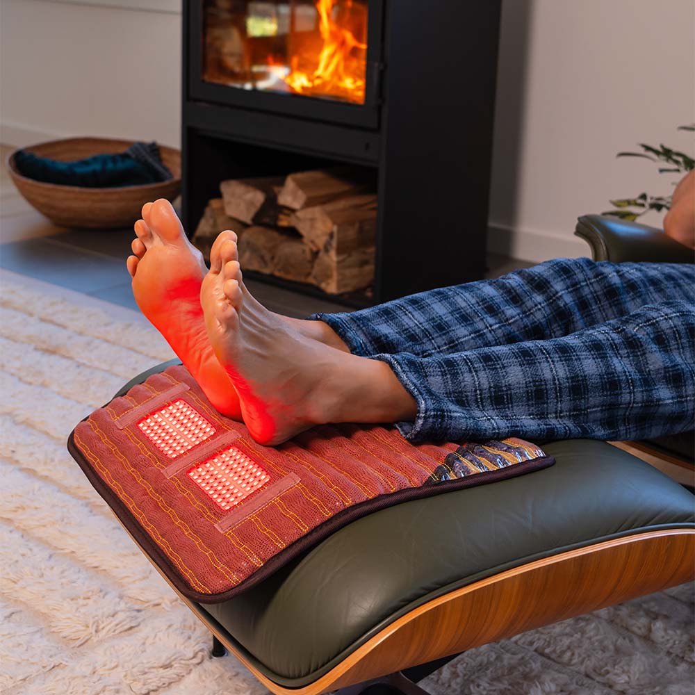 Bare feet on a HealthyLine TAO 18x18 Matrix mat with active red LED panels on a lounge ottoman, beside a fireplace