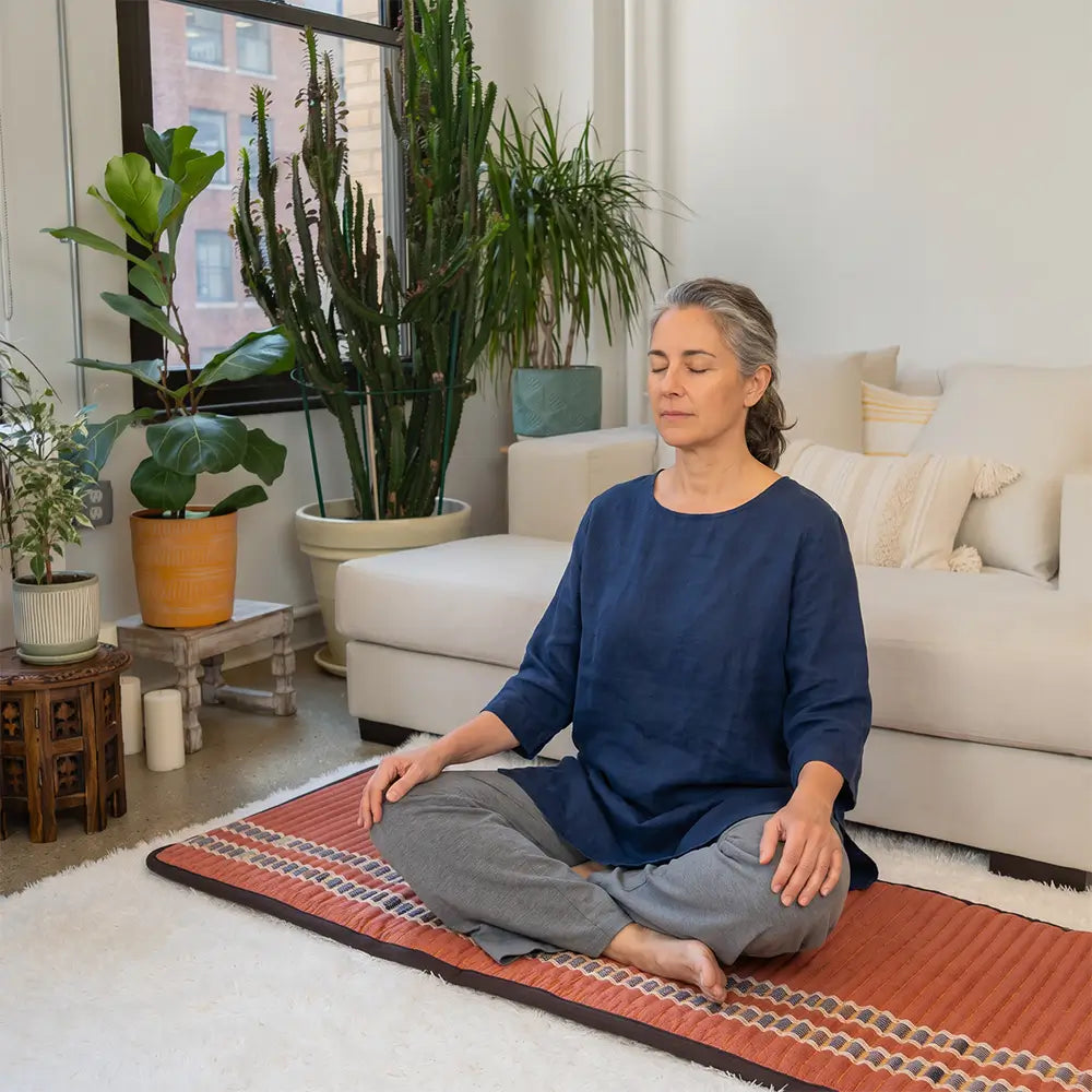 Woman meditates cross-legged with eyes closed on the red HealthyLine TAO Mat 7224, laid on a rug in a living room
