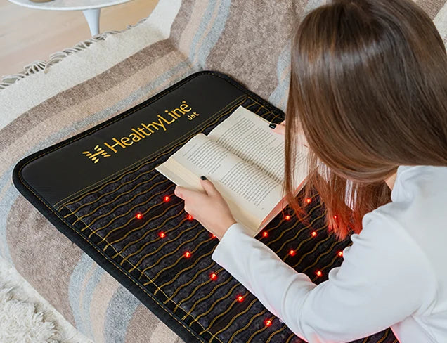 Woman reading a book while seated on a HealthyLine JET 72x24 mat with active red infrared LEDs on a living room floor