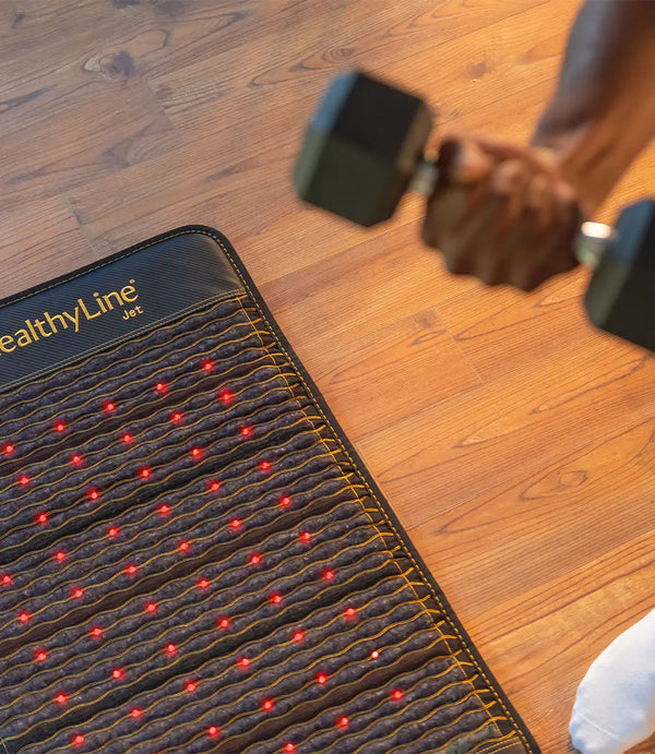 A person lifting a dumbbell next to a HealthyLine Jet 4024 mat with active red photon light therapy.