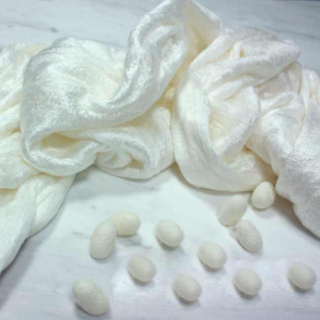 A draped bundle of raw white silk fibers next to silkworm cocoons on a marble surface.
